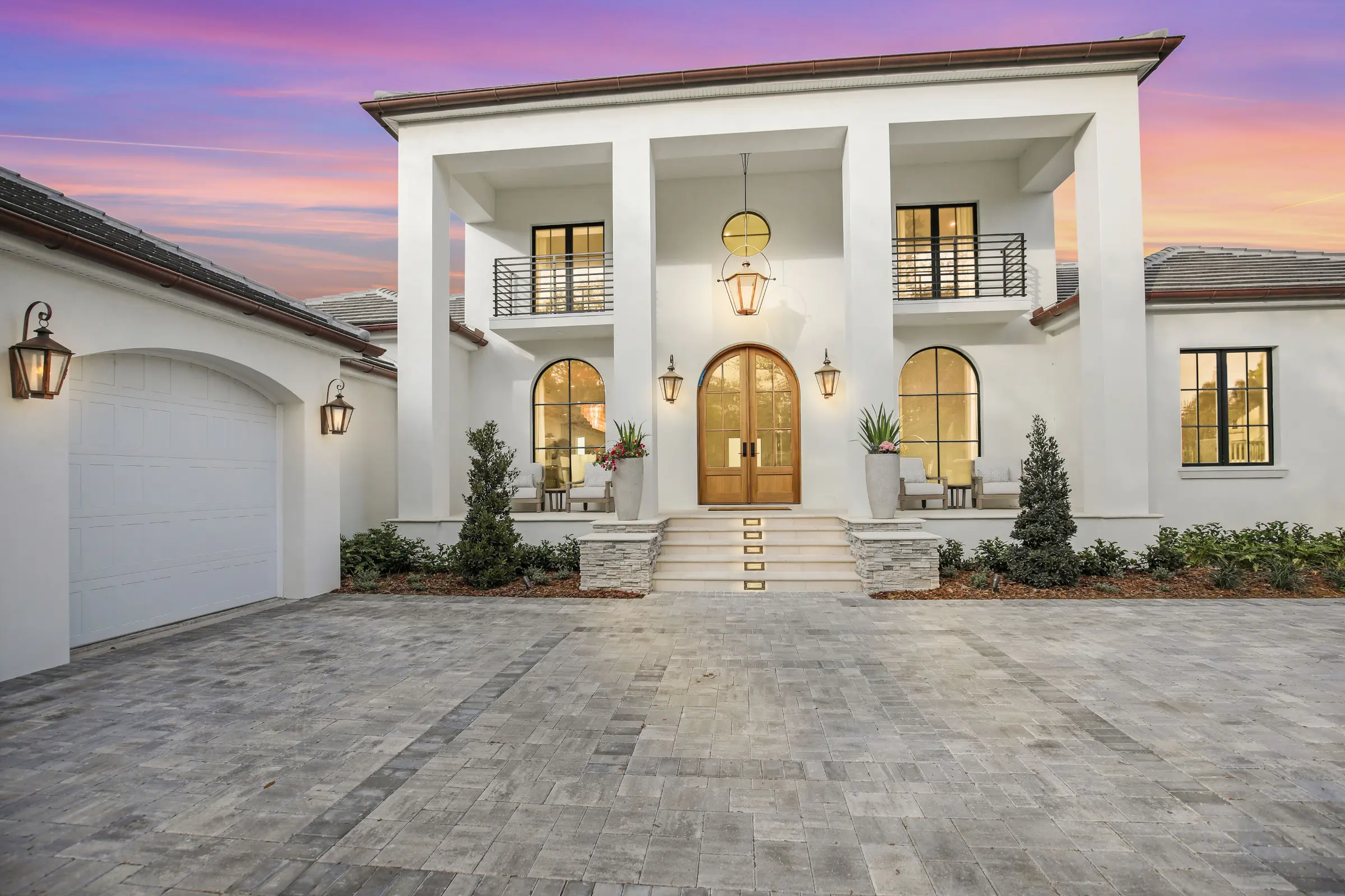 Modern white stucco home with arched entrance, illuminated windows, and paver driveway at sunset in Saint Petersburg, FL.