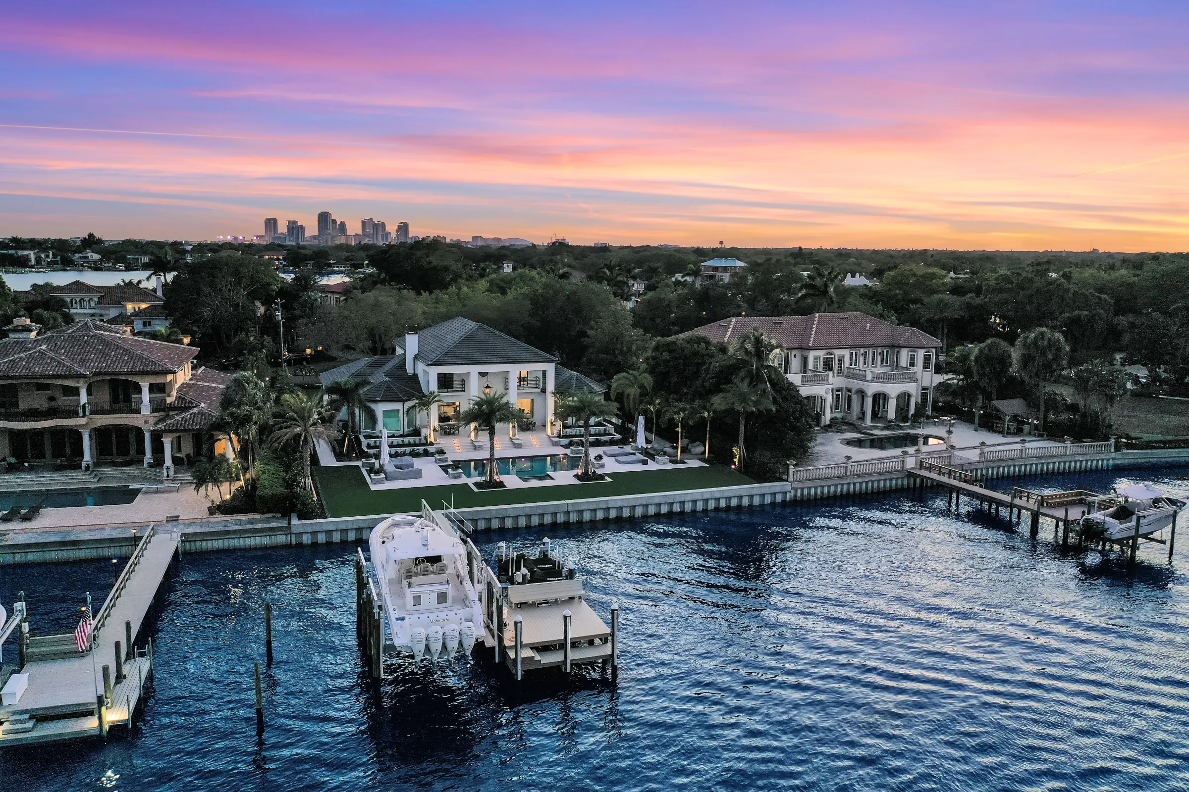 Luxury waterfront home with pool and boat dock at dusk, St Petersburg skyline visible in distance.