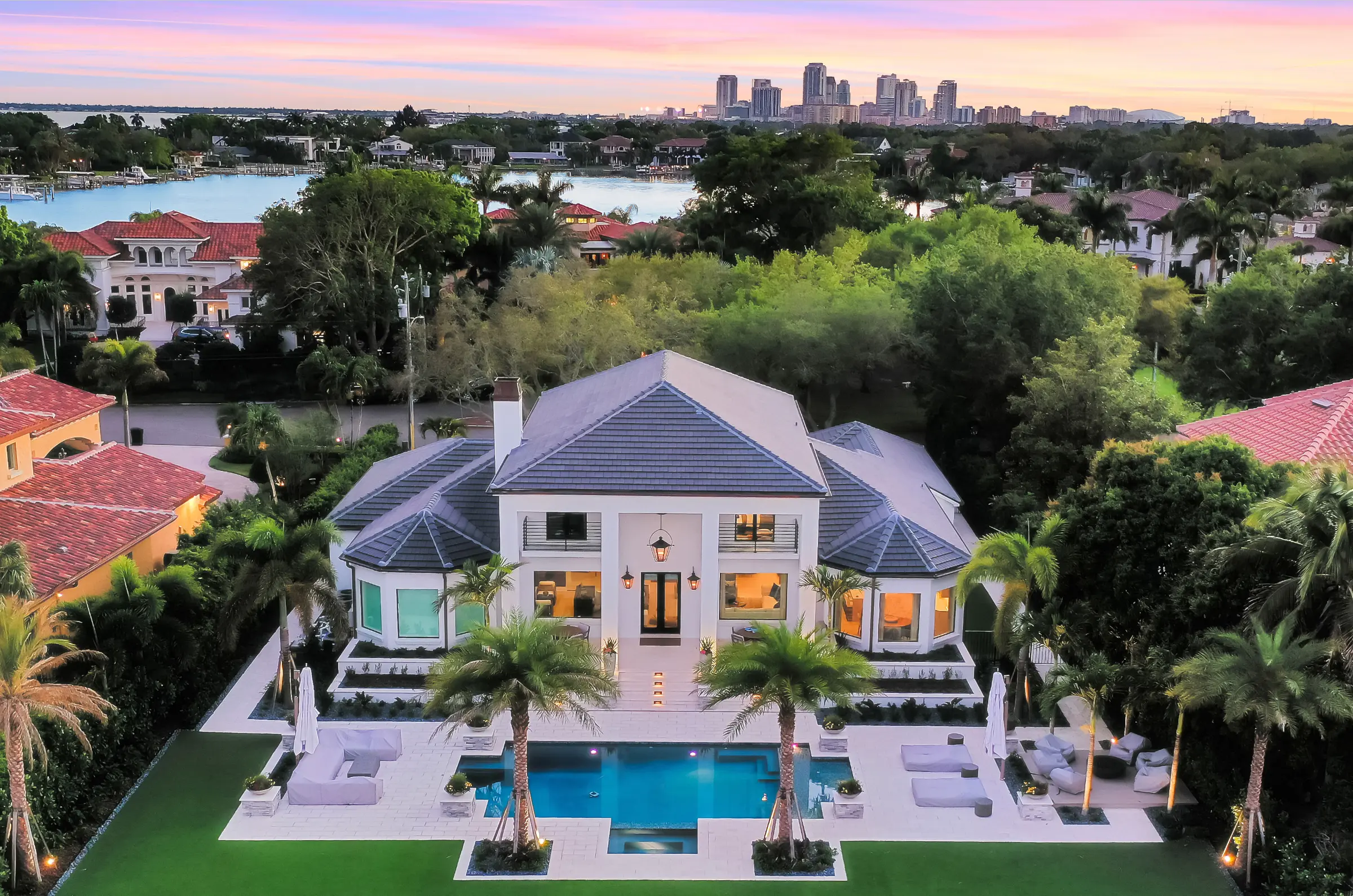 Luxury white contemporary home with blue roof and illuminated pool deck overlooking waterfront, exemplifying premium pool design and landscape architecture in St Petersburg, FL.