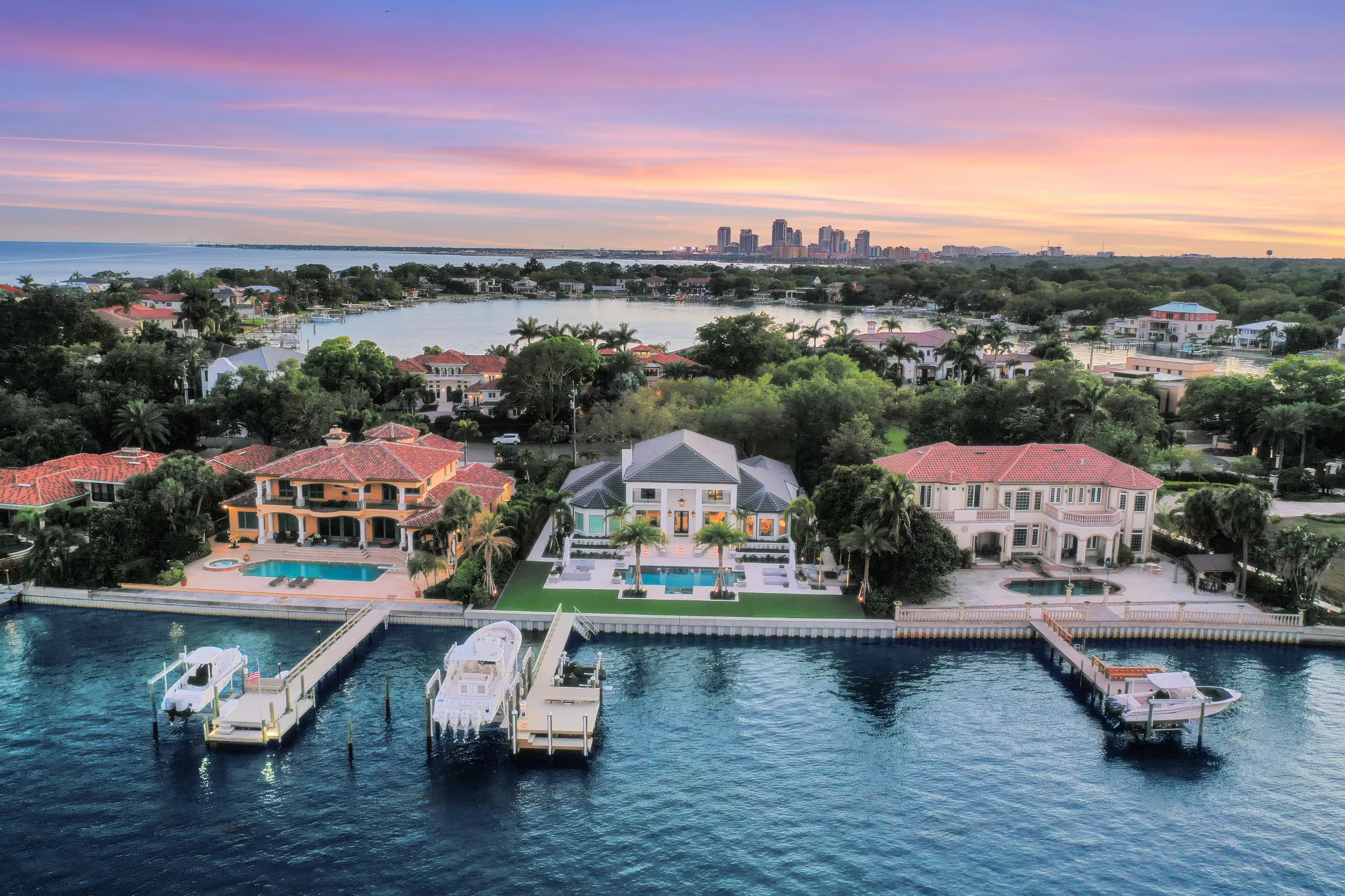 Luxury waterfront homes with pools and private boat docks overlook bay with distant city skyline at dusk in St Petersburg, FL.