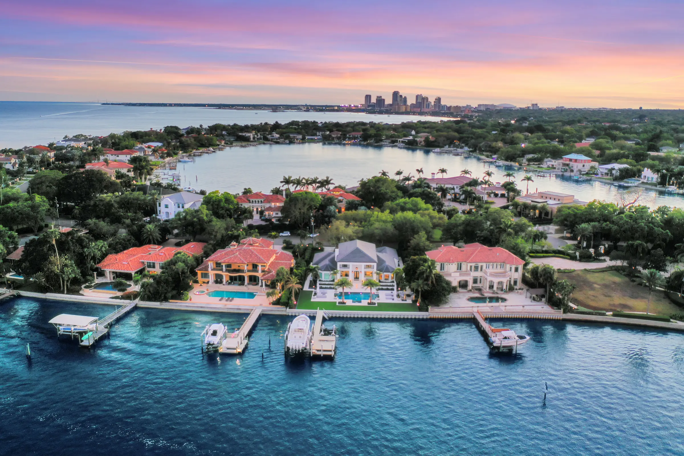 Luxury waterfront homes with pools and boat docks overlooking Biscayne Bay toward downtown Miami skyline at sunset