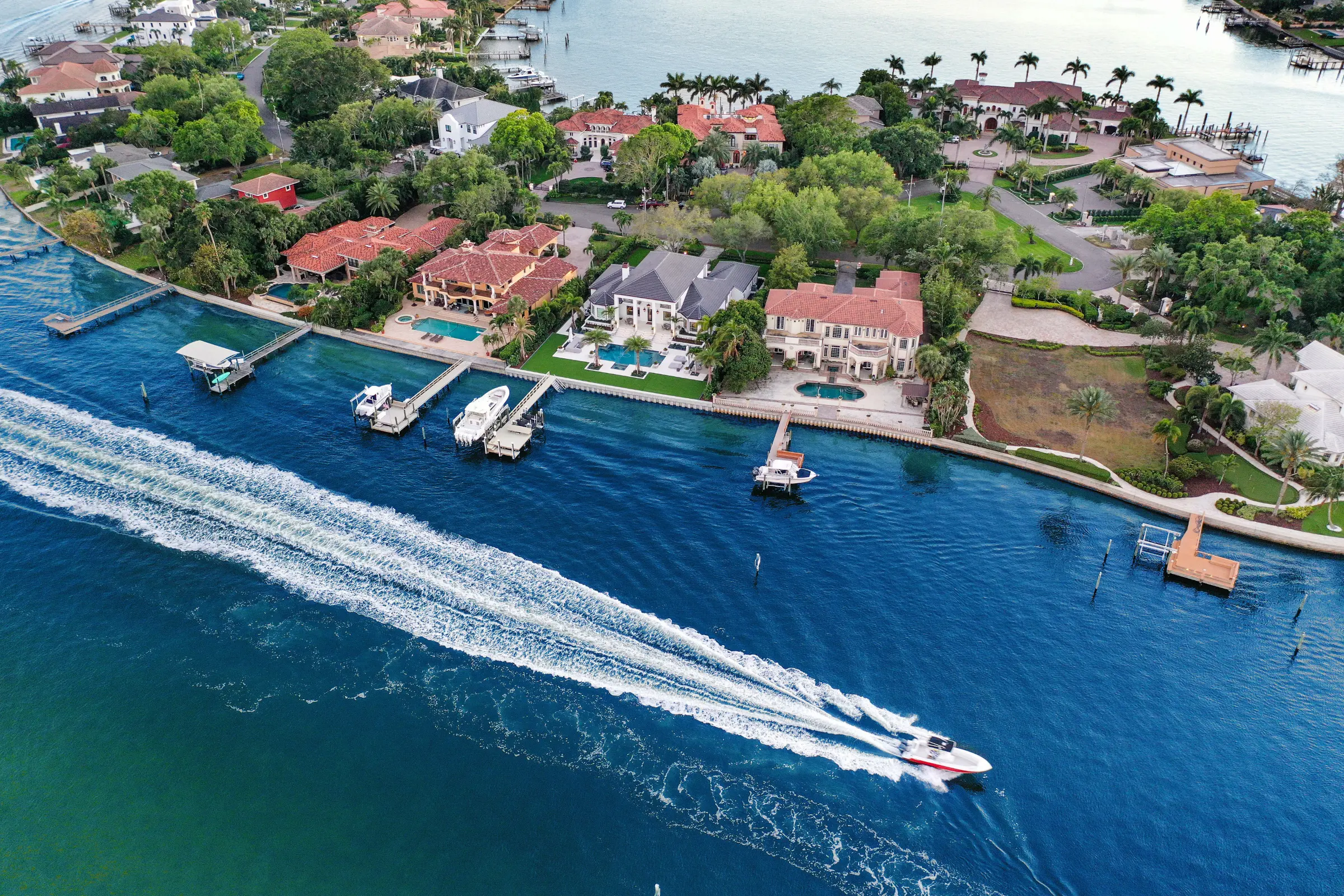 Aerial view of waterfront luxury homes with pools in St Petersburg, FL, featuring a speedboat racing through blue waters offshore.