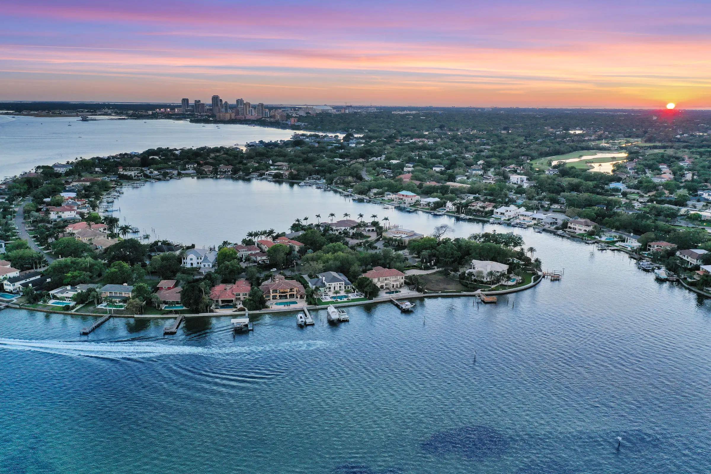 Aerial sunset view of luxury waterfront residential community with boats, pools, and distant city skyline across bay waters.