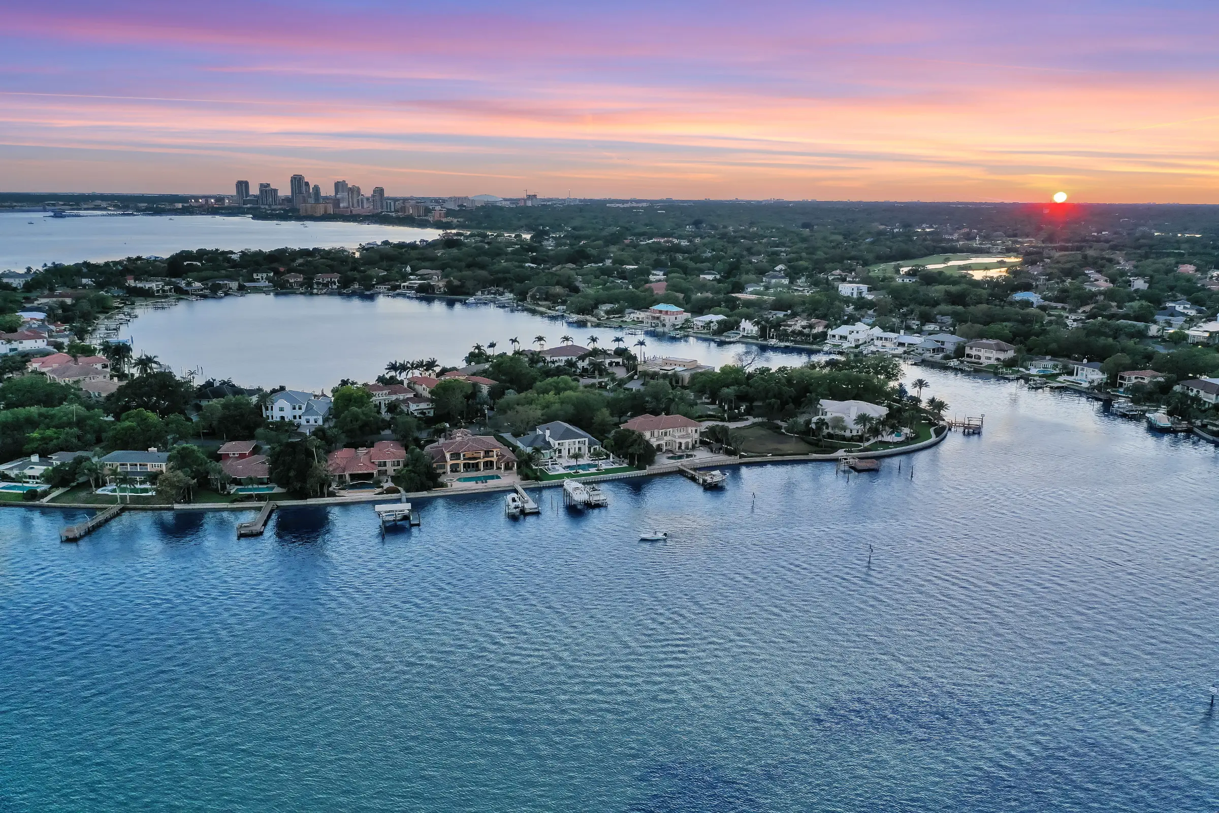 Aerial view of luxury waterfront homes with boats docked along canals at sunset, St Petersburg FL skyline in background.